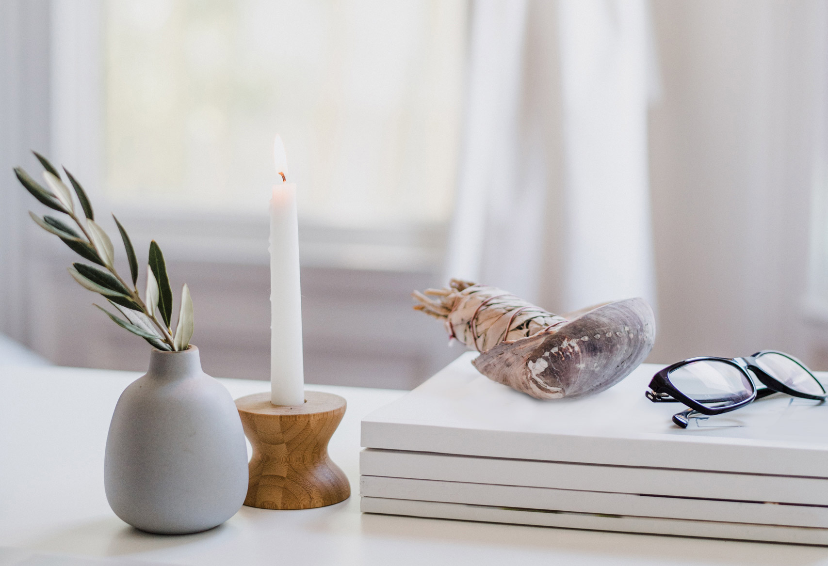A calm workspace with an olive branch in a vase, a lit candle, a stack of books and reading glasses